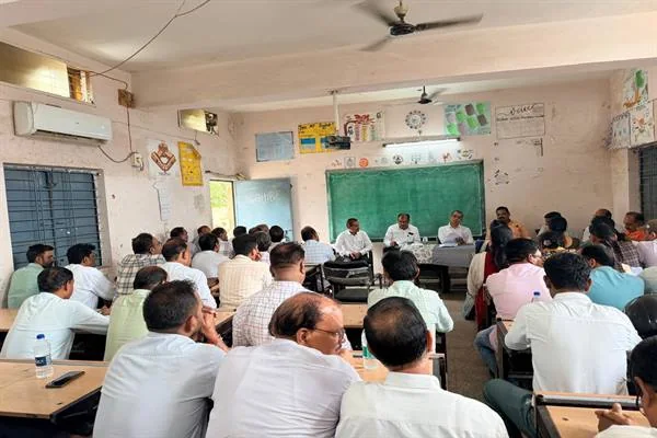 कक्षा में मेज-कुर्सियों पर बैठे लोगों के सामने मंच पर अधिकारी बैठक करते हुए