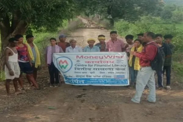 ग्रामीण रास्ते पर वित्तीय साक्षरता का बैनर थामे खड़े लोग और उन्हें जानकारी देता एक व्यक्ति