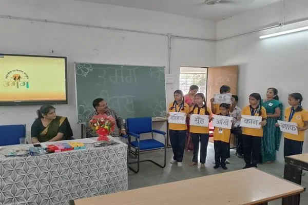 कक्षा में छात्राएं और विद्यार्थी हाथों में हिंदी शब्दों वाली तख्तियां लेकर खड़े हैं, सामने मेज पर दो महिलाएं बैठी हैं